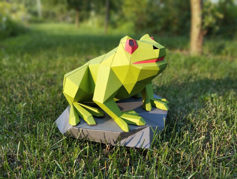 May include: A green origami frog with red eyes sitting on a gray rock. The frog is made of paper and has a geometric design.