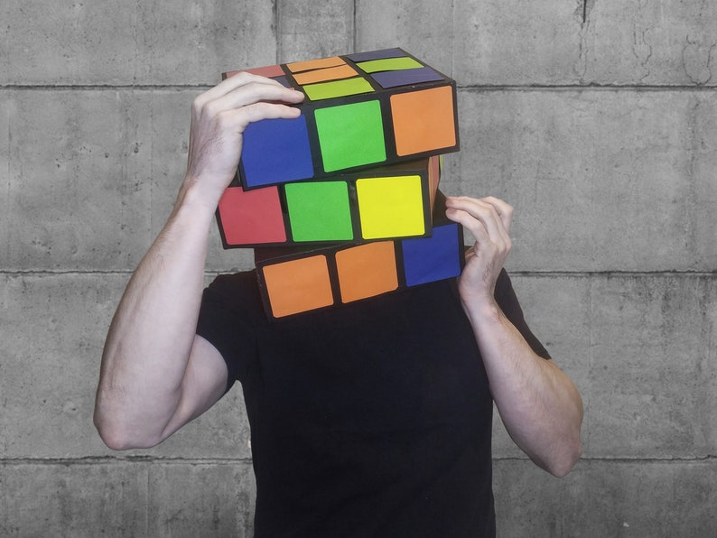 Pode incluir: Uma pessoa usando uma grande caixa de estilo cubo de Rubik colorida sobre a cabe&ccedil;a. A caixa &eacute; feita de papel&atilde;o e tem uma moldura preta com quadrados coloridos.