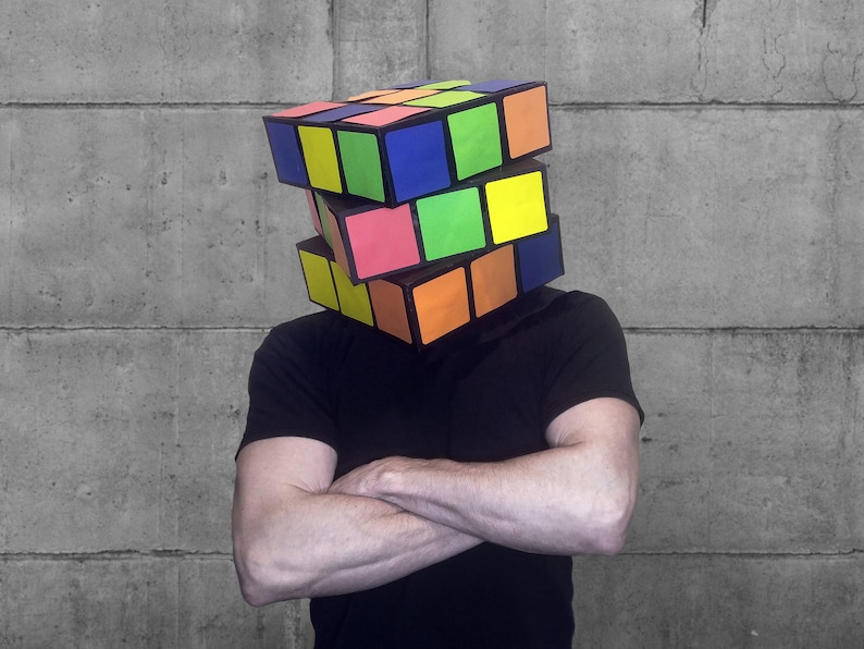 Pode incluir: Uma pessoa usando um grande cubo de Rubik colorido na cabe&ccedil;a. O cubo &eacute; feito de pl&aacute;stico e tem seis lados, cada um com uma cor diferente. A pessoa est&aacute; usando uma camisa preta e tem os bra&ccedil;os cruzados.