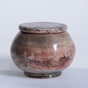 Puede incluir: Un tarro de cerámica hecho a mano con tapa. El tarro es redondo con una paleta de colores terrosos moteados de rosa, marrón y gris. La tapa refleja la forma y el color del tarro, creando un diseño cohesivo. El tarro se asienta sobre una pequeña base circular.