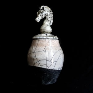 May include: A ceramic jar with a white and black glaze and a horse head lid. The jar has a crackled glaze and a distressed look.