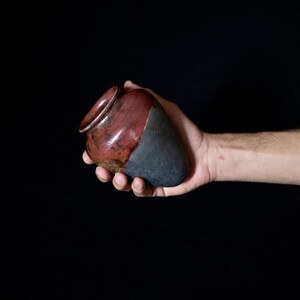 Könnte beinhalten: Eine kleine, handgefertigte Keramikvase mit einer dunkelbraunen und schwarzen Glasur. Die Vase hat die Form eines Herzens und einen abgerundeten Boden.