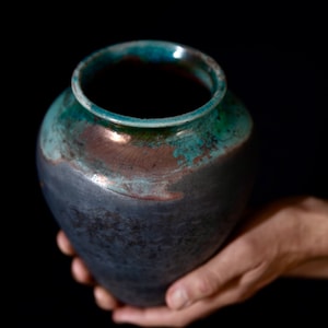 Könnte beinhalten: Eine dunkelgraue Keramikvase mit einer teal- und kupferfarbenen Glasur. Die Vase hat einen runden Körper und eine breite Öffnung.