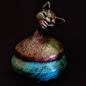 Puede incluir: Un tarro de cerámica con forma de gato con tapa. La base es de color azul turquesa y la tapa es multicolor. La cara del gato está pintada con una expresión caprichosa.