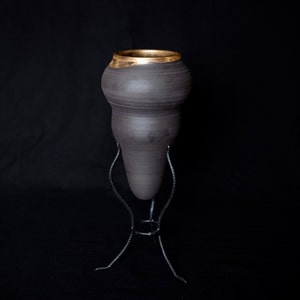 Könnte beinhalten: Eine schwarze Keramikvase mit goldfarbenem Rand, die auf einem schwarzen Metallständer mit drei Beinen steht.