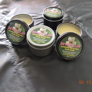 Op de afbeelding: Vier kleine zilveren blikken met Devil's Claw Herbal Healing Pain Rub. De blikken zijn gelabeld met een witte achtergrond en zwarte tekst. De tekst luidt "Devil's Claw Herbal Healing Pain Rub. May help lower back pain & joint inflammation. All Natural Ingredients. 4 oz."