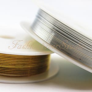 May include: Two spools of wire, one gold and one silver, are wound around white spools. The spools are on a white surface.