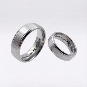 May include: Two matching silver wedding rings. Each ring has a brushed metal finish and a polished edge. The inscription "Forever Love 05/01/2020" is engraved on the inside of each ring. The rings are displayed on a white background.