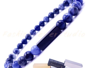 Puede incluir: Un brazalete de cuentas de piedras azules con un dije rectangular negro y dos dijes más pequeños grabados con "06.08.2022" y "Forever".