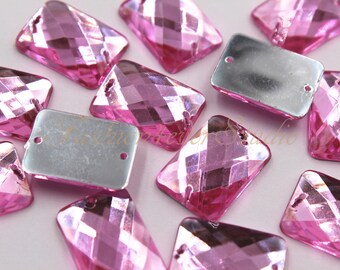 May include: Close-up of several pink rectangular crystal beads with faceted surfaces. Each bead has a silver backing with two holes for stringing. The beads are scattered, showcasing their reflective qualities.
