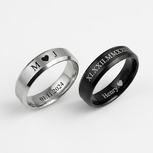 May include: Two rings: one silver, the other black. The silver ring is engraved with the initials "M" and "J", a heart symbol, and the date "01.11.2024". The black ring features Roman numerals and the name "Henry" with a heart. Both rings have a brushed metal finish.