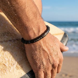 Peut inclure: Bracelet noir avec coordonnées gravées sur une plaque métallique noire. Le bracelet est porté sur un bras bronzé tenant une planche de surf sur une plage de sable. L'océan est visible en arrière-plan.