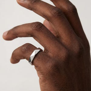 Può includere: Un anello geometrico argentato con un design sfaccettato. L'anello ha una forma moderna e angolare con molteplici superfici piatte che riflettono la luce. L'anello è mostrato su un dito.