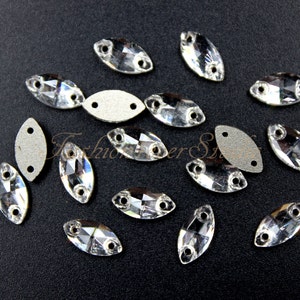 May include: A collection of clear, faceted, marquise-shaped rhinestones with two holes each. The rhinestones are scattered on a black surface.