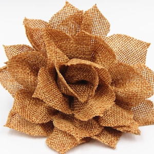 May include: A large, handcrafted flower made from woven burlap fabric. The flower has multiple layers of petals, creating a full, textured appearance. The burlap is a warm, natural brown color, and the flower is set against a white background.