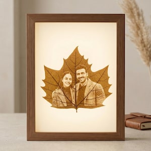 May include: A framed, illuminated artwork featuring a couple's portrait etched into a large, brown leaf. The wooden frame surrounds the leaf, which is lit from behind, creating a warm glow. The couple is smiling.
