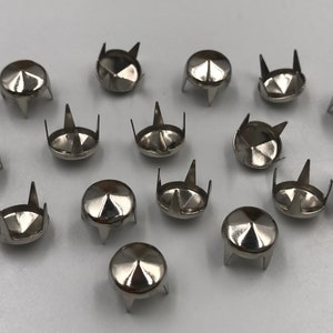 Könnte beinhalten: Eine Sammlung silberfarbener Metallpyramidennieten mit drei Zacken an der Unterseite. Die Nieten sind auf einer weißen Oberfläche verstreut.
