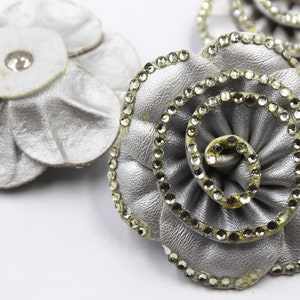 May include: A silver leather flower with a rhinestone trim. The flower has a layered design with a center swirl.