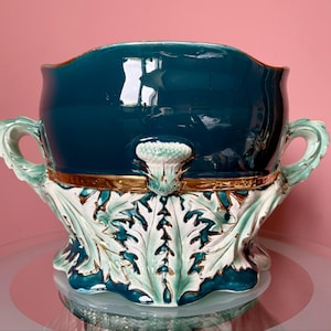 Op de afbeelding: Een grote, teal-kleurige keramische plantenbak met een gouden rand en witte en groene bloemdetails. De plantenbak heeft twee handvatten en een brede, ronde basis.