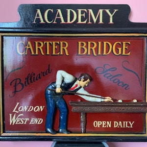Peut inclure: Une enseigne en bois vintage pour le Carter Bridge Billiard Saloon dans le West End de Londres. L'enseigne présente une image 3D d'un homme jouant au billard, avec les mots "Academy", "Billiard Saloon", "London West End", et "Open Daily" en lettres dorées.