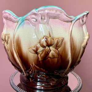 Op de afbeelding: Een vintage keramische plantenpot met een gegolfde rand en een bloemenmotief. De pot is versierd met bruine, roze en groene glazuur en heeft een reliëf bloemenmotief.