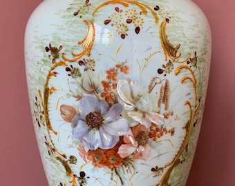 Puede incluir: Un jarrón grande de cerámica blanca con un diseño floral verde y dorado. El jarrón tiene una boca ancha y una base estrecha. El diseño floral presenta un ramo de flores blancas, rosas y naranjas con detalles dorados.