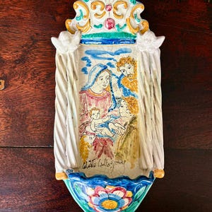 Op de afbeelding: Een handbeschilderde keramische wanddecoratie met een religieuze scène. Het stuk heeft een wit, blauw, geel en roze kleurenschema. De onderkant heeft een bloemenontwerp en de bovenkant een decoratieve kroon. Het kunstwerk toont een familie.