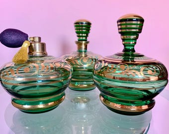 Puede incluir: Tres botellas de perfume vintage de vidrio con detalles dorados y vidrio verde. Las botellas tienen diferentes formas y tamaños.