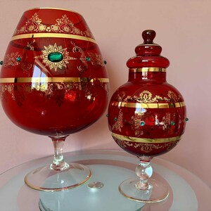 Op de afbeelding: Twee sierlijke, rode glazen decoratiestukken. Het grotere stuk is kelkvormig en het kleinere is een pot met deksel. Beide hebben gouden filigraanontwerpen, smaragdgroene accenten en gouden banden. De stukken staan op heldere glazen bases.