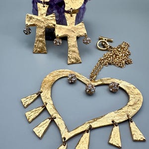 Op de afbeelding: Goudkleurige sieradenset met een hartvormige hangerketting met bungelende accenten, een ketting en kruisvormige oorbellen. De oorbellen hebben hartvormige toppen en bungelende kristallen accenten. De sieraden hebben een getextureerde afwerking.