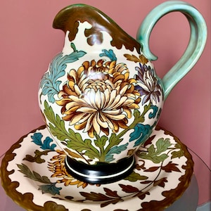 Op de afbeelding: Een witte keramische kan met een groene handgreep en een bijpassend bord. Beide stukken zijn versierd met een bloemmotief van bruine, gele en blauwe chrysanten.