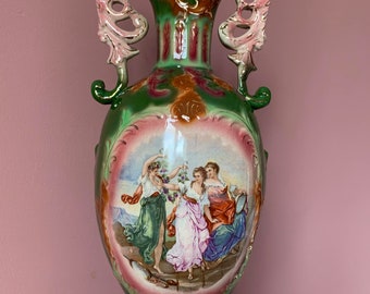 May include: A large, green and brown ceramic vase with ornate handles and a painted scene of three women in a landscape. The vase has a pink and white floral design.
