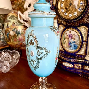 Puede incluir: Un tarro de cristal decorativo azul claro con tapa, con filigranas doradas y pequeños detalles rojos. El tarro tiene una base de cristal transparente y un pomo turquesa en la parte superior. El tarro está sobre una superficie de madera.