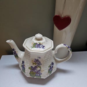 Puede incluir: Una tetera de cerámica blanca con un diseño floral de violetas moradas. La tetera tiene un patrón de celosía y una tapa con un pequeño pomo. Un jarrón blanco alto con una decoración de corazón rojo está en el fondo.