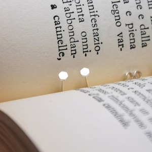 May include: Two silver stud earrings with a hexagonal shape, placed on the pages of an open book.