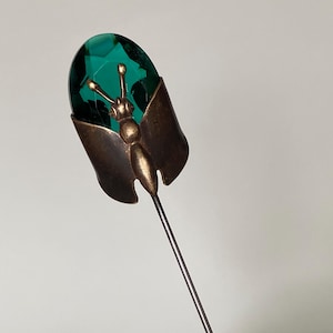 Könnte beinhalten: Eine Anstecknadel mit einem bronzefarbenen Metallschmetterling mit ausgebreiteten Flügeln. Der Schmetterlingskörper ist auf einem facettierten, ovalen smaragdgrünen Edelstein zentriert, der den oberen Teil des Insektendesigns bildet.