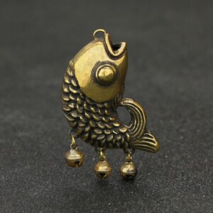 Könnte beinhalten: Ein goldfarbener Messingfisch-Anhänger mit einem detaillierten Design und drei kleinen Glocken, die an der Unterseite hängen.