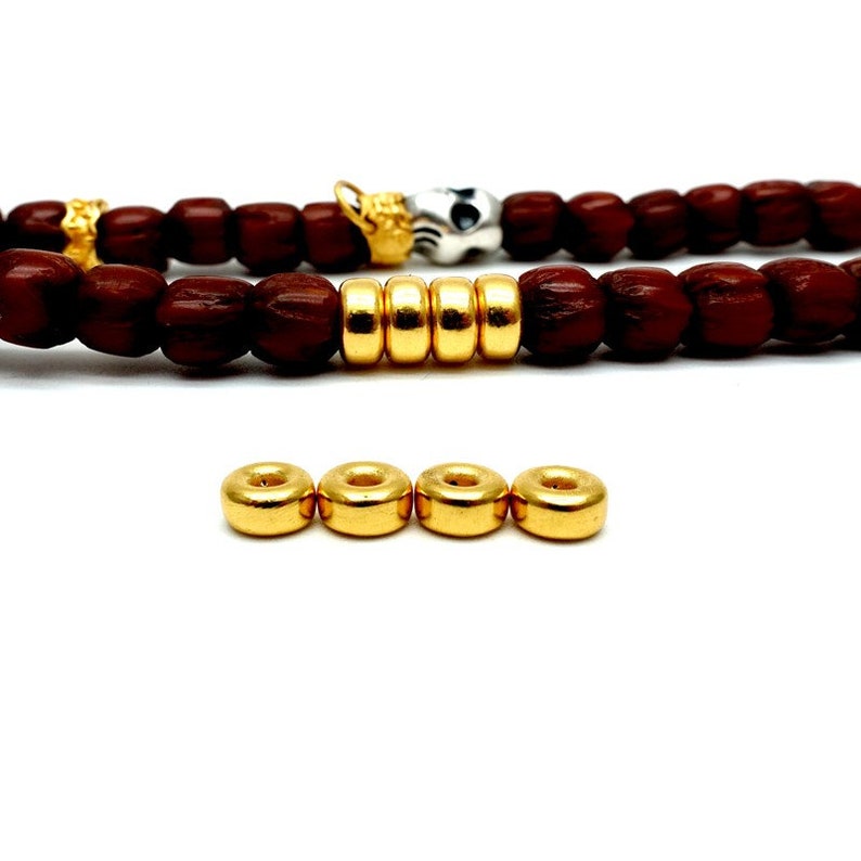 May include: A close-up of four gold-colored metal beads. The beads are round and have a hole in the center. The beads are arranged in a row on a white surface.