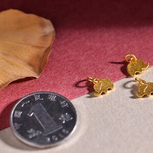 May include: Small gold-colored elephant charms with a loop for jewelry making. The charms are displayed with a coin and a dried leaf on a neutral background. The elephant charms are approximately 0.5 inches in size.