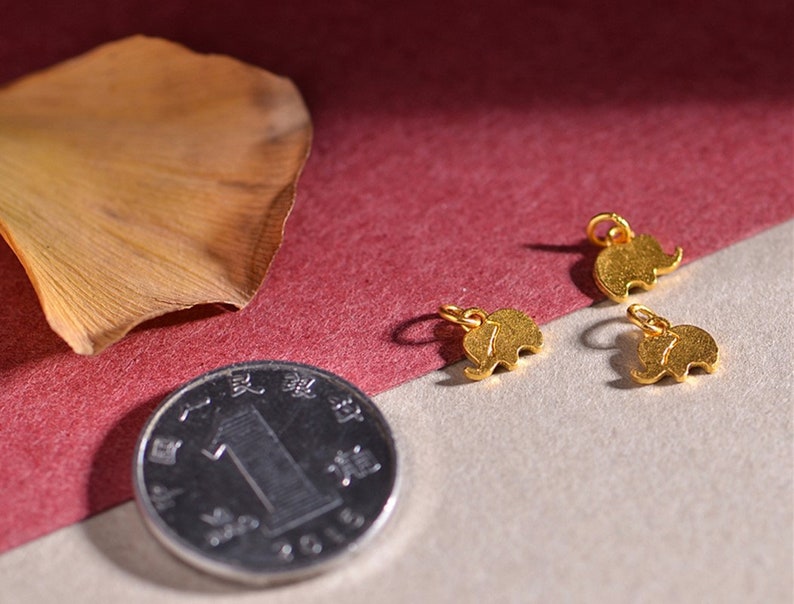 May include: Small gold-colored elephant charms with a loop for jewelry making. The charms are displayed with a coin and a dried leaf on a neutral background. The elephant charms are approximately 0.5 inches in size.