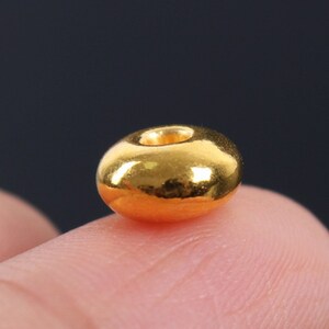 May include: A close-up of a single, shiny, gold-colored bead. The bead is round with a hole through the center. The bead is resting on a finger, with a dark background.