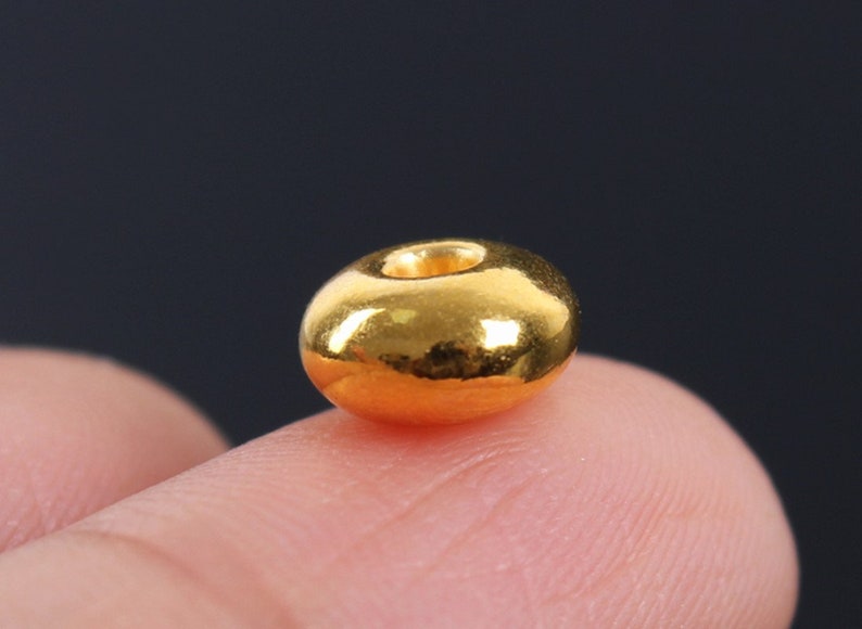 May include: A close-up of a single, shiny, gold-colored bead. The bead is round with a hole through the center. The bead is resting on a finger, with a dark background.