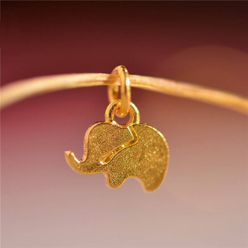May include: A gold-colored elephant charm pendant. The charm is a silhouette of an elephant with textured details. The pendant is suspended from a thin, gold-colored chain, against a blurred background.