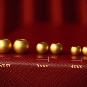K&ouml;nnte beinhalten: Sechs goldfarbene Perlen in verschiedenen Gr&ouml;&szlig;en von 4 mm bis 6 mm Durchmesser sind in einer Reihe auf einer roten Oberfl&auml;che angeordnet. Die Perlen sind glatt und gl&auml;nzend.