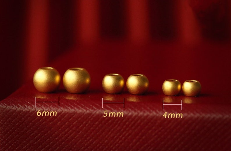 K&ouml;nnte beinhalten: Sechs goldfarbene Perlen in verschiedenen Gr&ouml;&szlig;en von 4 mm bis 6 mm Durchmesser sind in einer Reihe auf einer roten Oberfl&auml;che angeordnet. Die Perlen sind glatt und gl&auml;nzend.