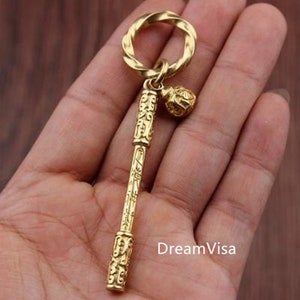 May include: A gold-coloured keychain with an ornate design. It features a twisted ring, a decorative central piece, and a small, round charm. The design includes intricate patterns and symbols, with a detailed finish.