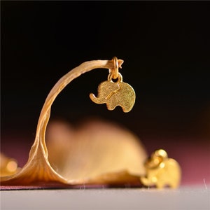May include: A gold-colored elephant charm pendant suspended from a thin, curved stem. The charm is detailed with etched lines and hangs against a blurred background. A second, smaller elephant charm is visible in the foreground.
