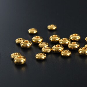 May include: A collection of small, shiny, gold-colored beads. Each bead has a rounded, donut-like shape with a hole through the center. The beads are scattered on a dark surface, reflecting light and creating highlights.