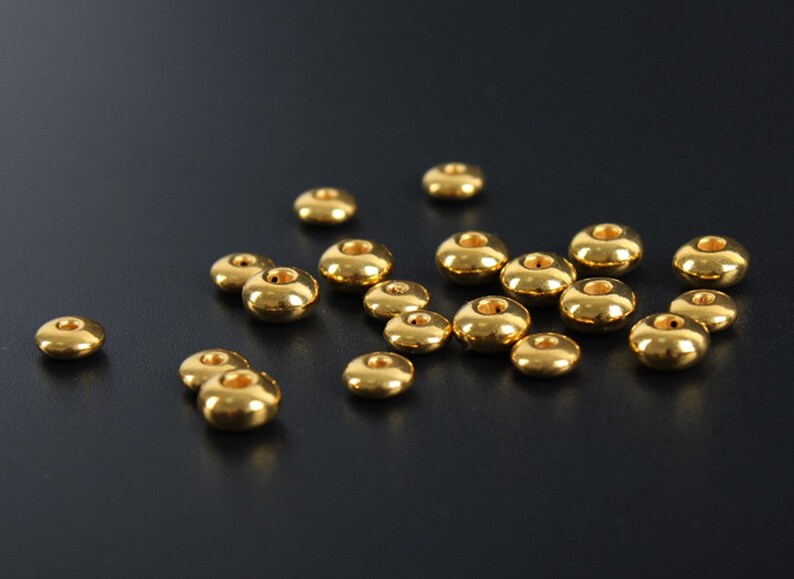 May include: A collection of small, shiny, gold-colored beads. Each bead has a rounded, donut-like shape with a hole through the center. The beads are scattered on a dark surface, reflecting light and creating highlights.