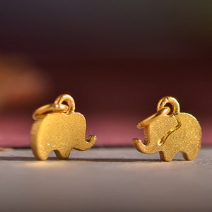 May include: Two small, gold-colored elephant charms with textured surfaces. Each charm has a loop at the top for attaching to a chain or earring hook. The elephant charms are displayed against a blurred, colorful background.